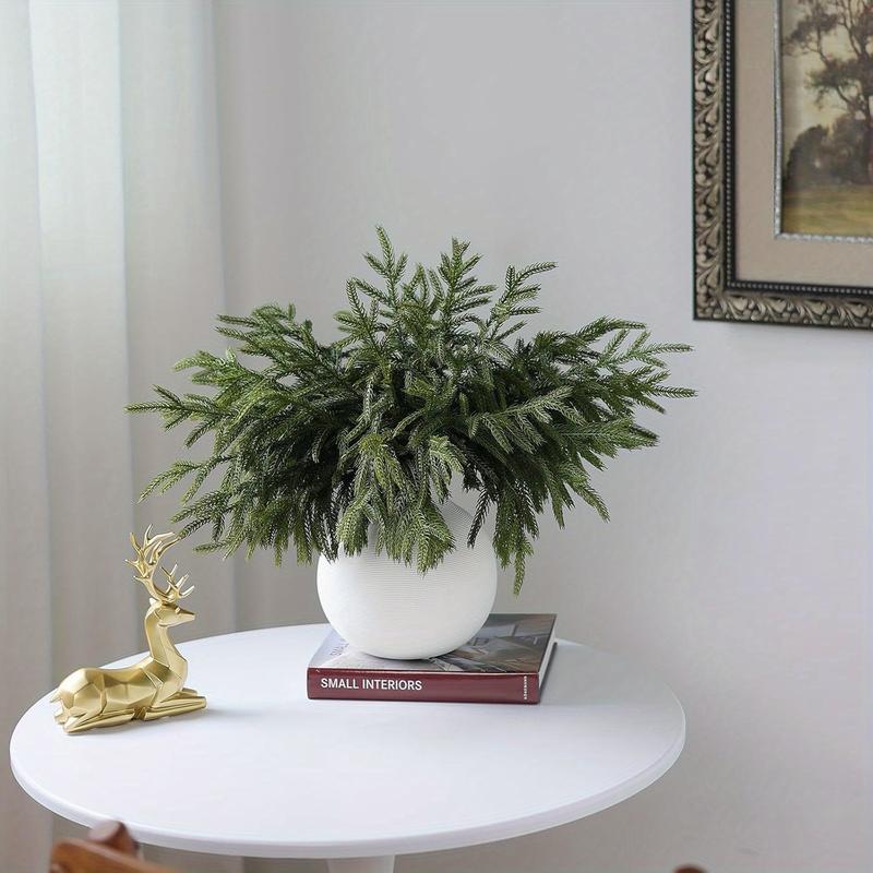 Green artificial Norfolk pine branches in a round white vase on books beside a gold holiday ornament