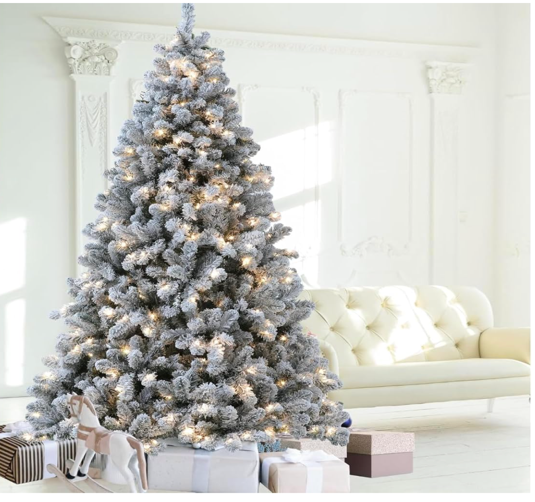 Silvanest snow-flocked Christmas tree with warm white lights beside a white sofa and wrapped gifts