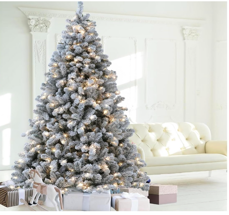 Snow-flocked Christmas tree with warm white lights and wrapped gifts beside a white sofa